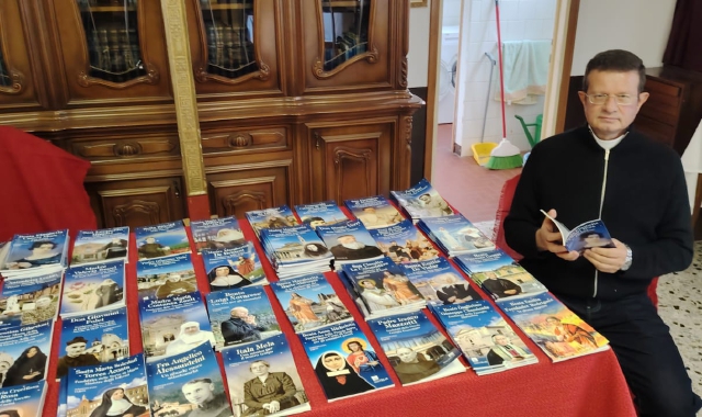 Don Massimiliano Taroni con tutti i   volumi (finora cento) che ha scritto  in questi anni su santi, martiri e altre figure eminenti della Chiesa Cattolica